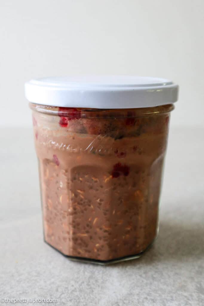 Shaken up old fashioned oats, chia seeds, cacao powder, honey, milk and topped with raspberries and cacao nibs, all in a jar, covered with white lid.