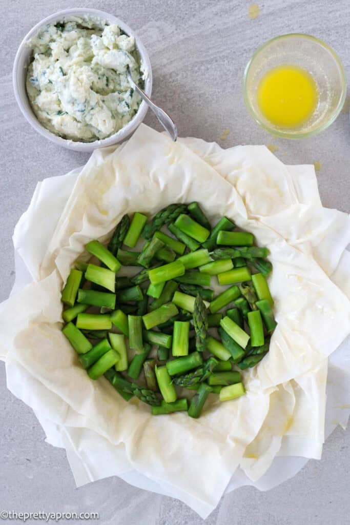 Asparagus in pie dish on top of phyllo dough and small bowl of ricotta cheese